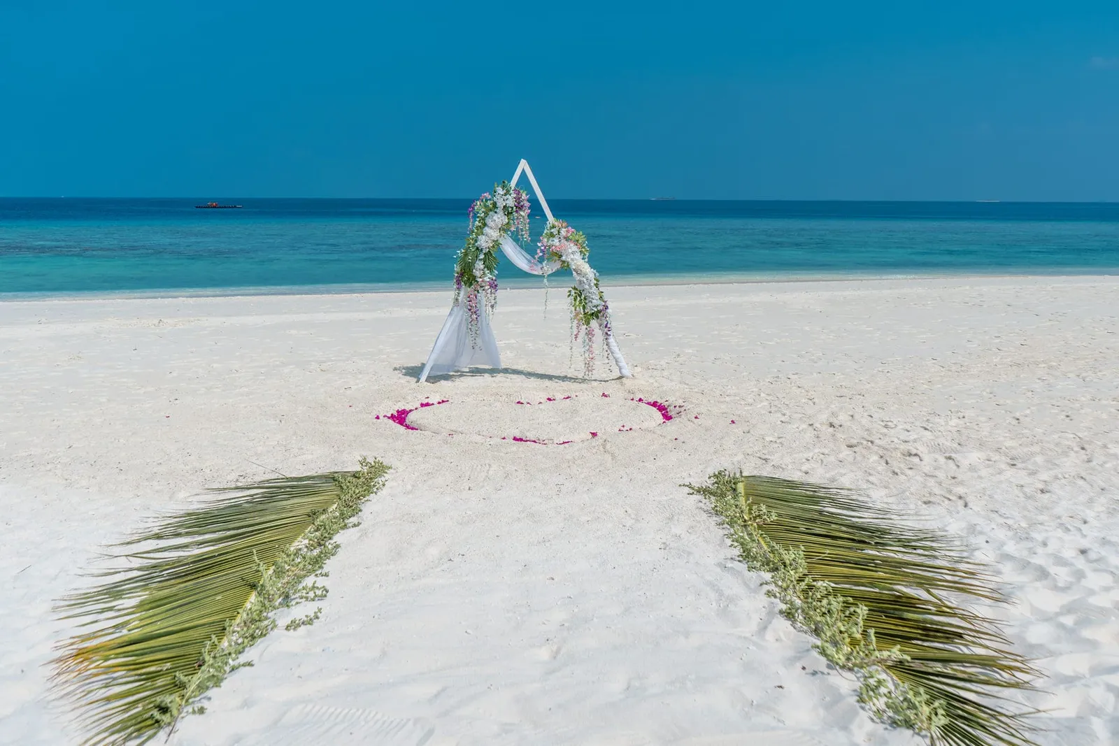 The wedding arch on the beach, with flowers, a heart of petals on the sand and 2 lines of palm fronds.