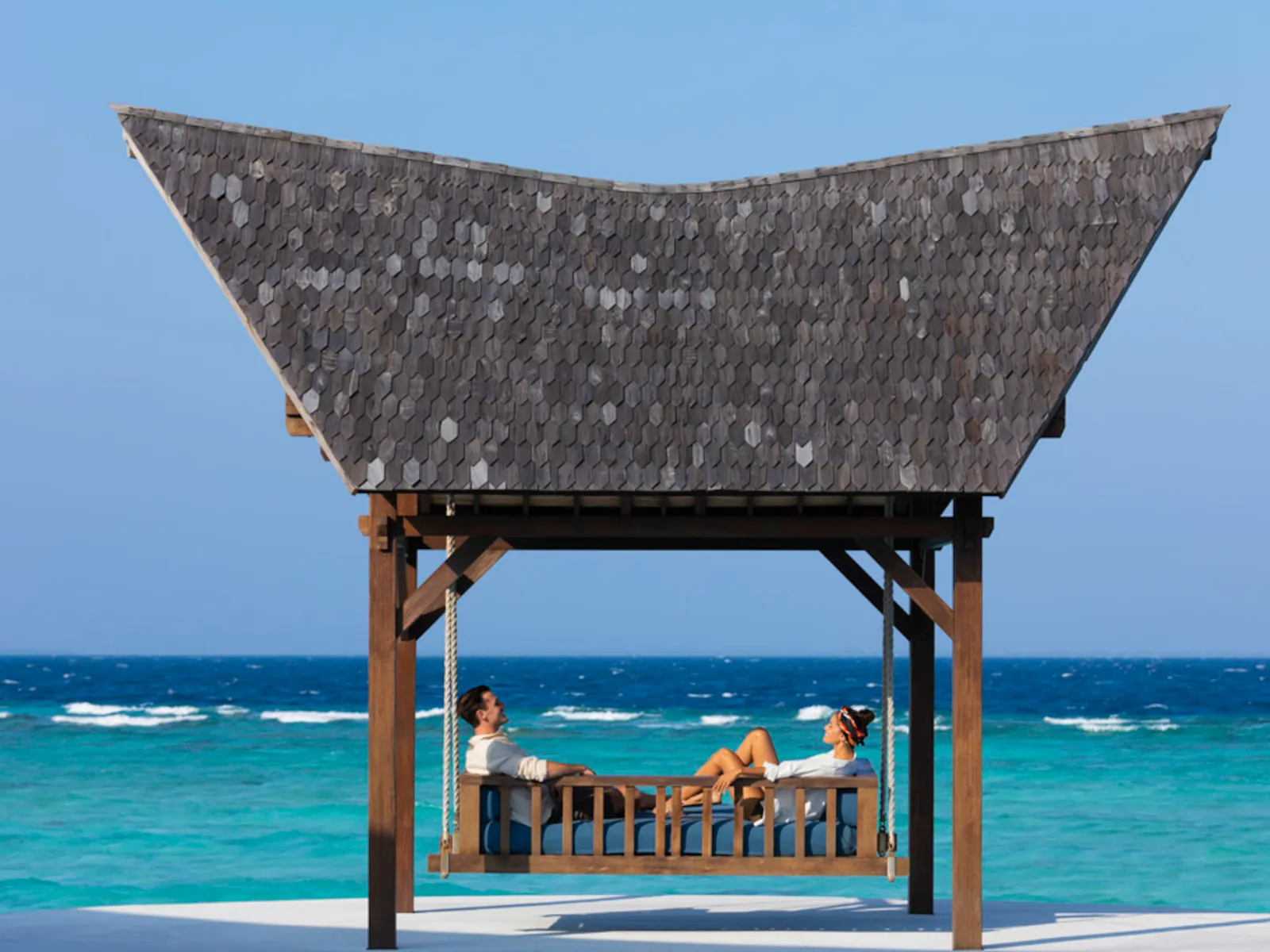 A swing under a thatched roof on the shore, a couple sitting before the sea