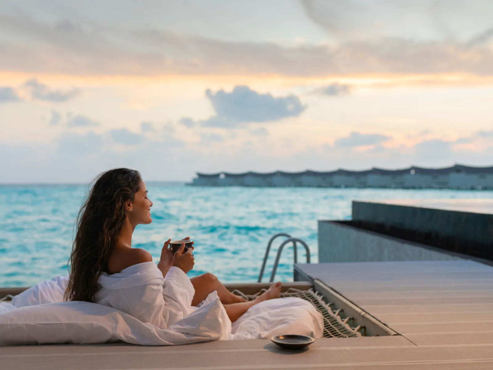 A woman on an overwater villa’s deck at sunrise, beside the pool