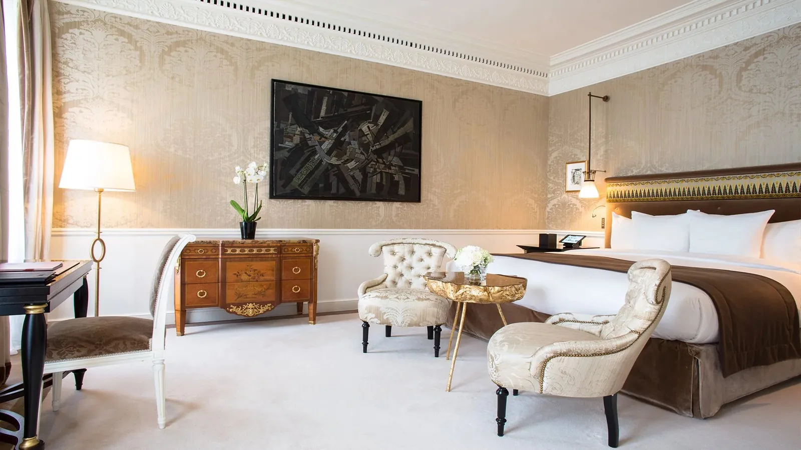 A bedroom with a bed, 2 pale armchairs and a table