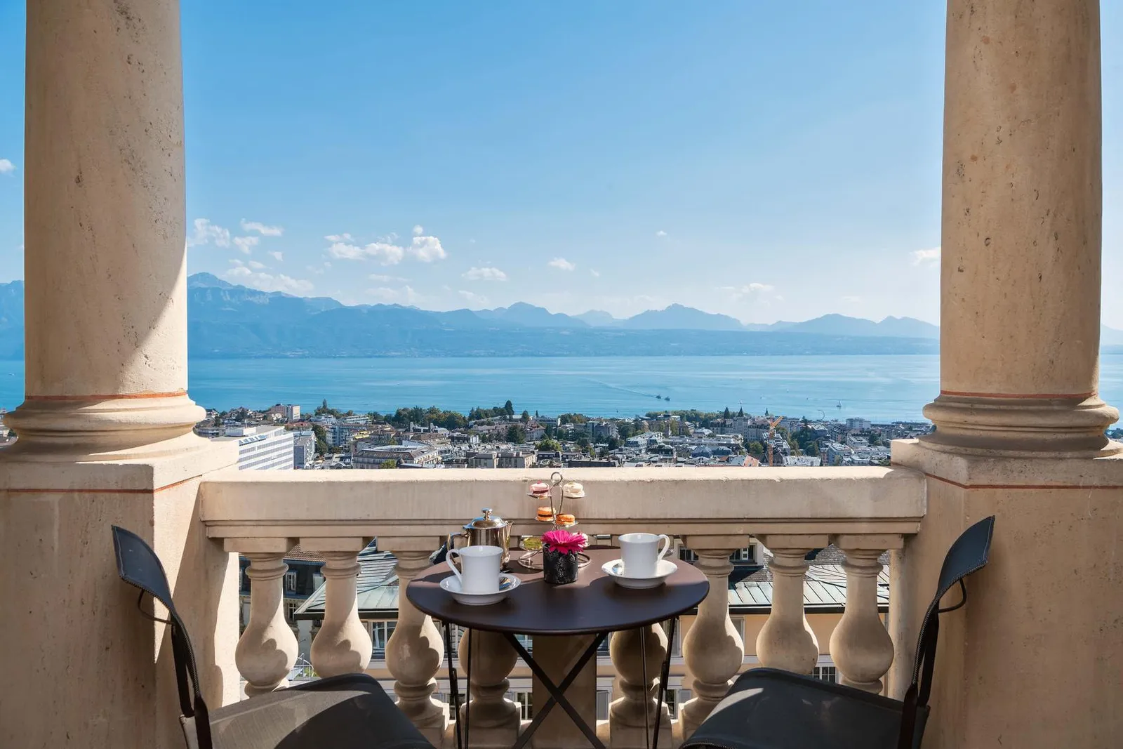 The balcony of a Suite Lake View between 2 stone columns: a small table set with tea and pastries and 2 chairs facing Lake Geneva, with the Alps on the far shore and the rooftops of Lausanne below