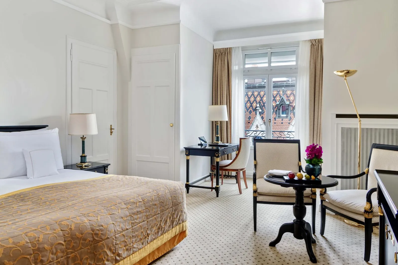 A Classic Business City Room: a bed with a gold bedspread, a small round table with 2 armchairs, a writing desk and a window onto a roof of patterned tiles