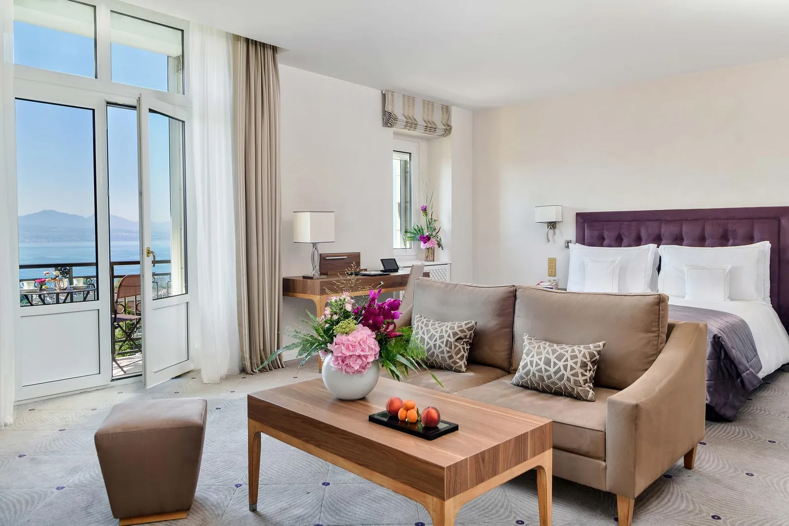 A Deluxe Room Lake View: a sofa, a coffee table with flowers and fruit, a bed with a purple headboard and the French window open onto the balcony, with the lake and mountains in the distance