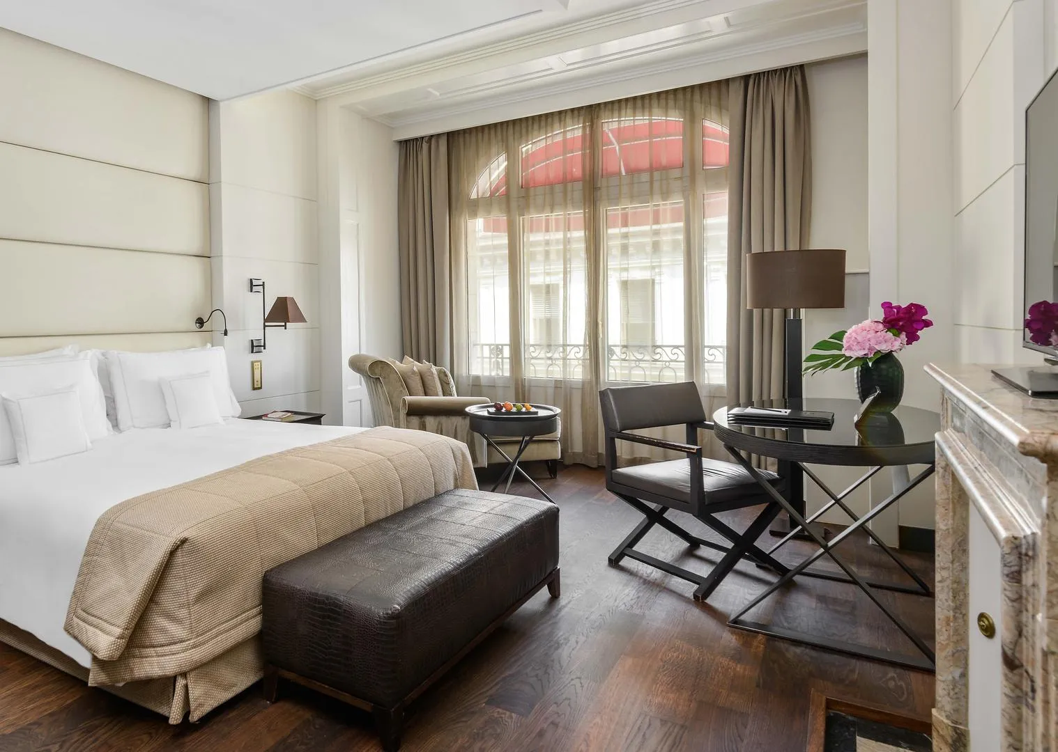 A Premium City Room: a bed with a padded headboard, a dark leather bench, parquet floor, a marble fireplace and an arched window with a red awning outside
