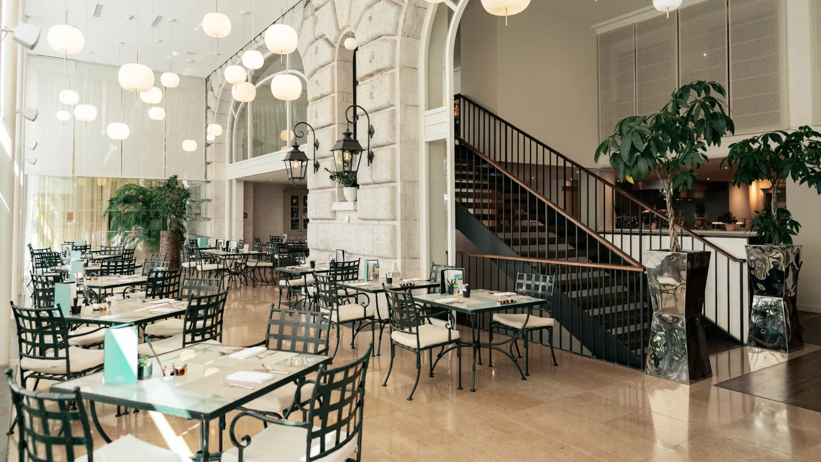 Côté Jardin, a winter-garden dining room: green iron tables and chairs with white cushions, globe lamps, a stone wall with lanterns, a staircase and large plants
