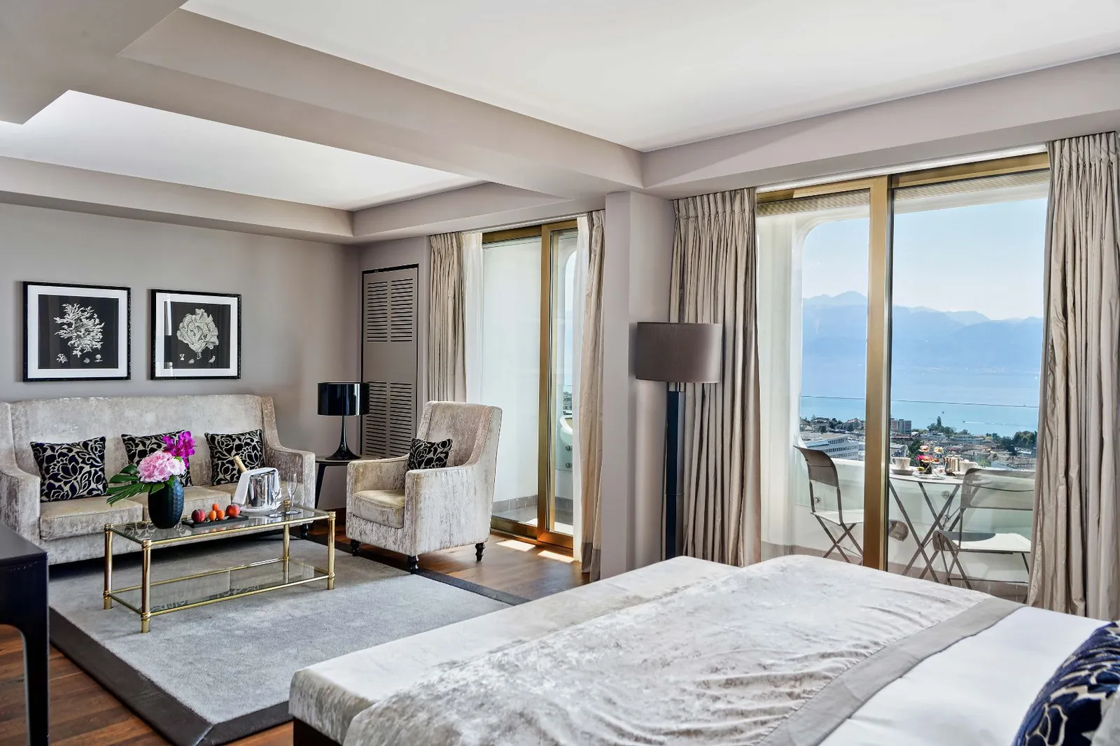 A Junior Suite Lake View: the bed in the foreground, a sitting area with a sofa and armchairs and French windows onto the balcony, with the lake and mountains beyond