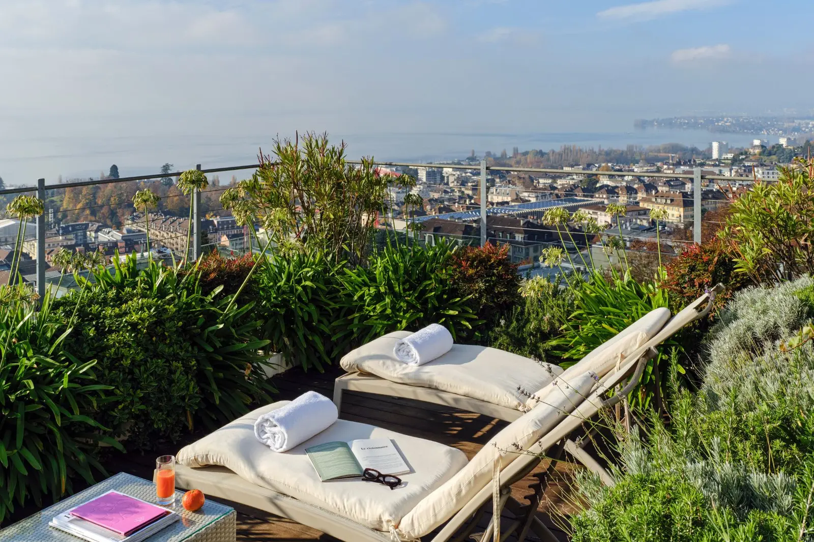 The Penthouse terrace: 2 loungers with towels among Mediterranean plants, a small table with magazines and a glass of juice, and below, the city’s rooftops running down to the lake