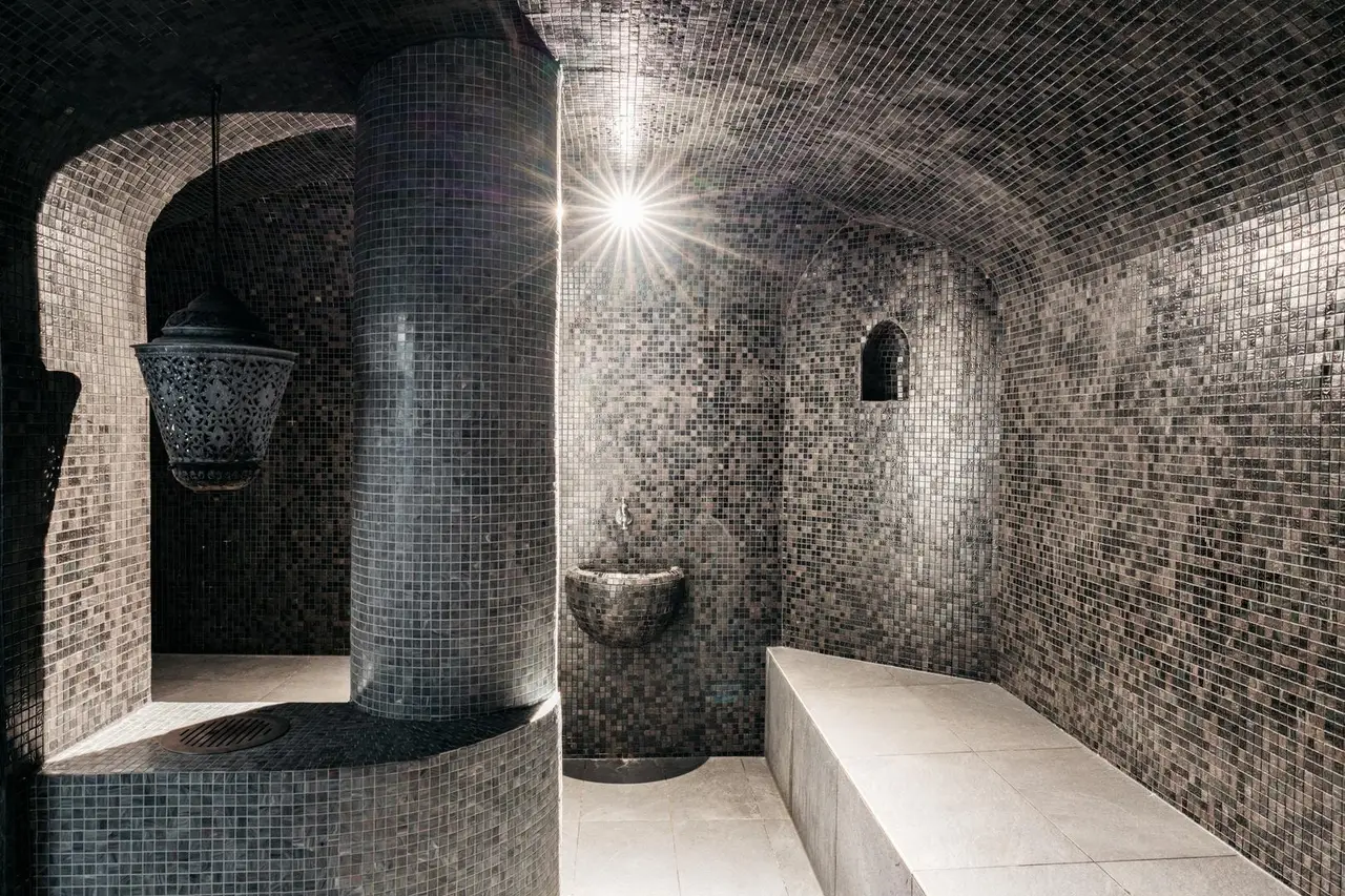 The hammam: walls and vault clad in small dark mosaic tiles, a mosaic column, a pierced lantern, a wall basin and a pale stone bench