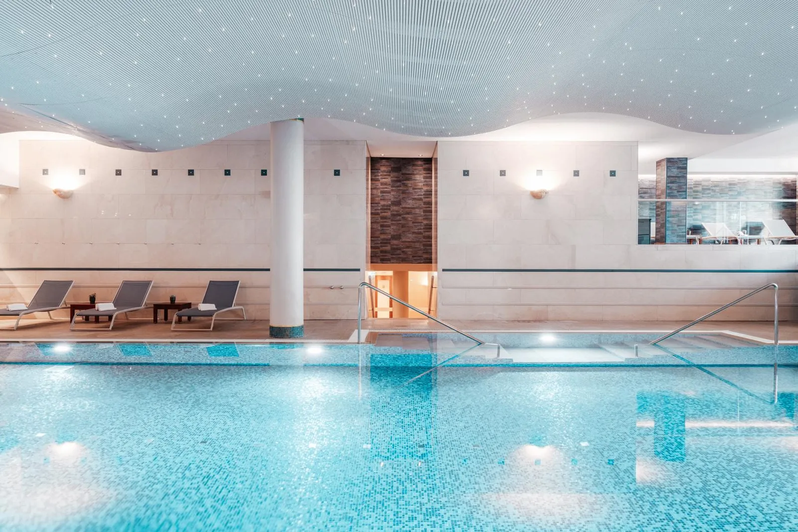 The indoor pool seen head-on: blue water over mosaic tiles, 3 loungers along the edge, a white column and the wave-shaped ceiling with small lights
