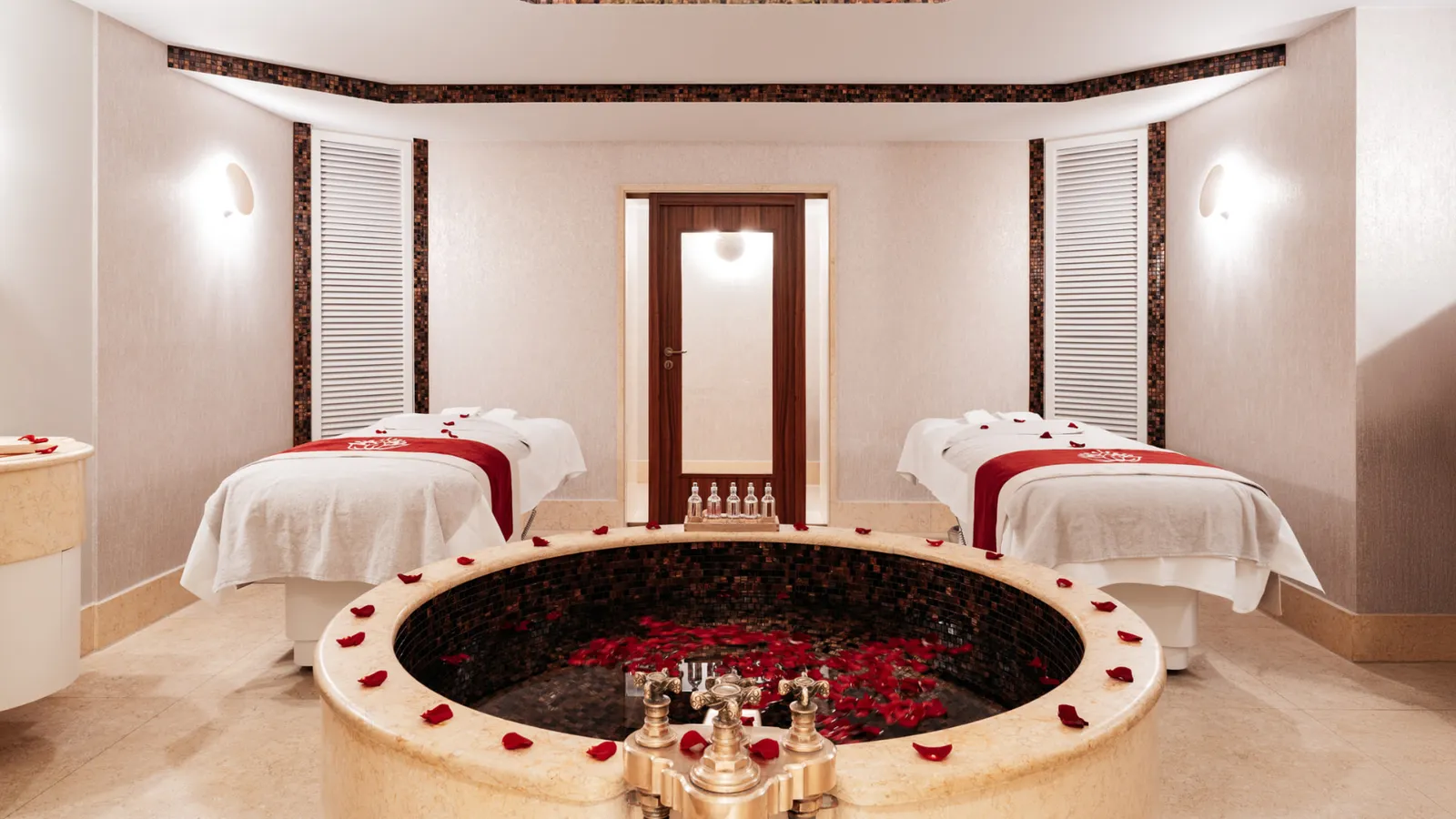 The private Spa Suite: 2 massage tables dressed in white and red either side of a round mosaic bath strewn with rose petals, with gold-coloured taps