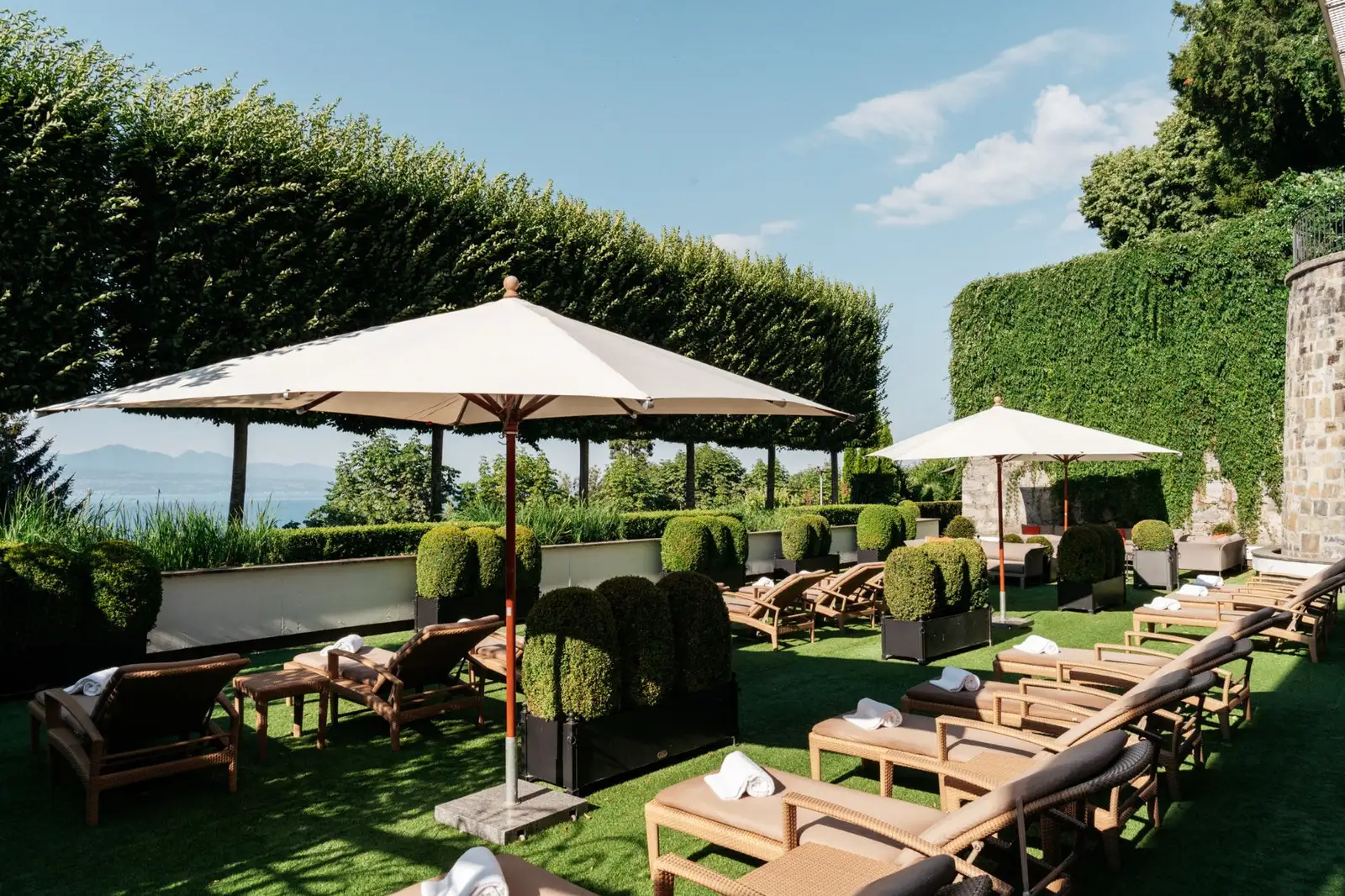 The spa’s garden terrace: loungers with towels on the lawn, box hedges in planters, 2 large white parasols and tall clipped hedges; a glimpse of the lake on the left