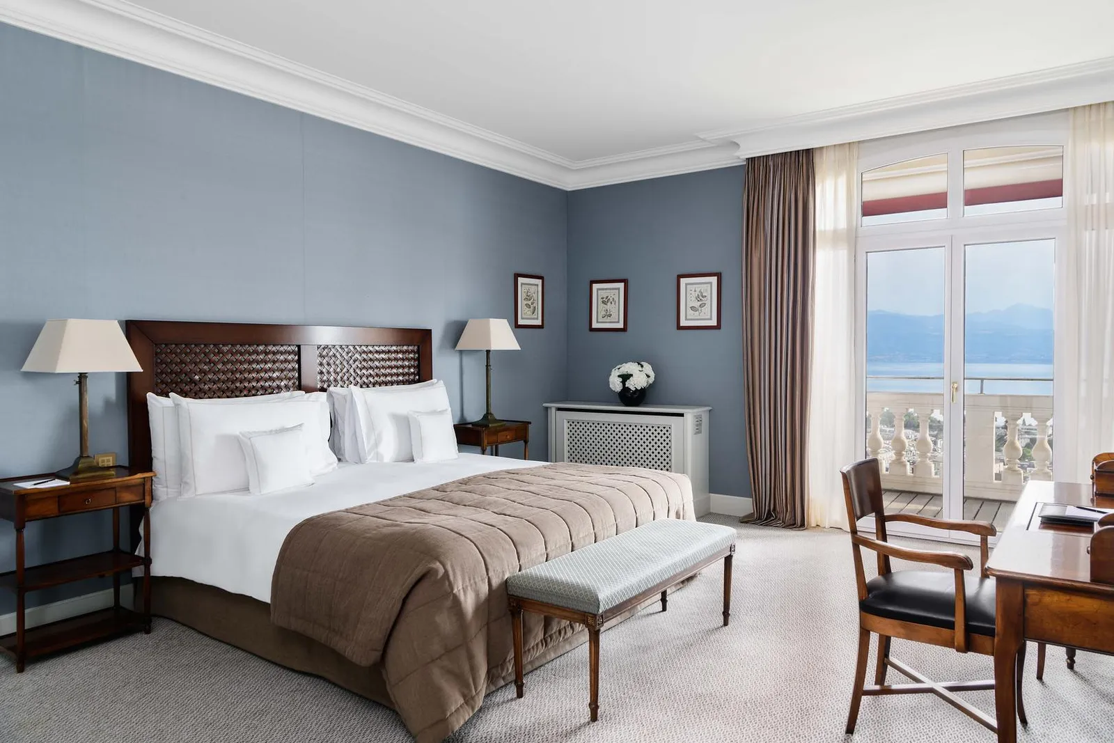 The bedroom of the Coco Chanel Suite: blue-grey walls, a bed with a carved wooden headboard, 2 shaded lamps and the French window onto a balustraded balcony overlooking the lake