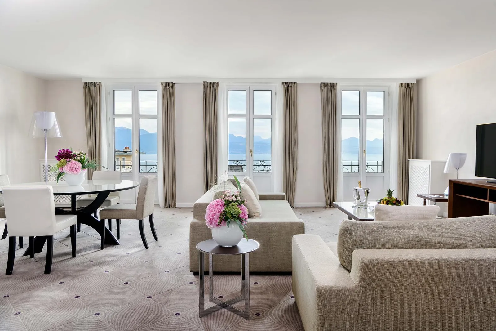 The living room of the Presidential Suite: pale sofas, a round dining table and 3 French windows looking out over the lake and the Alps