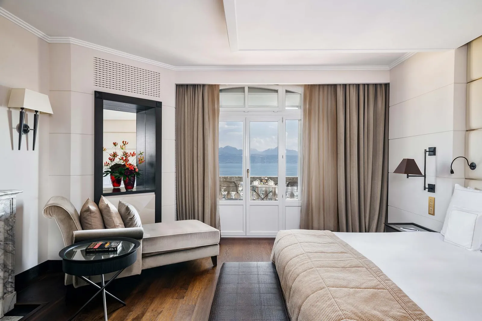 The bedroom of the Samaranch Suite: a bed with a sand-coloured bedspread, a chaise longue, dark parquet and the French window onto the balcony, with the lake and mountains