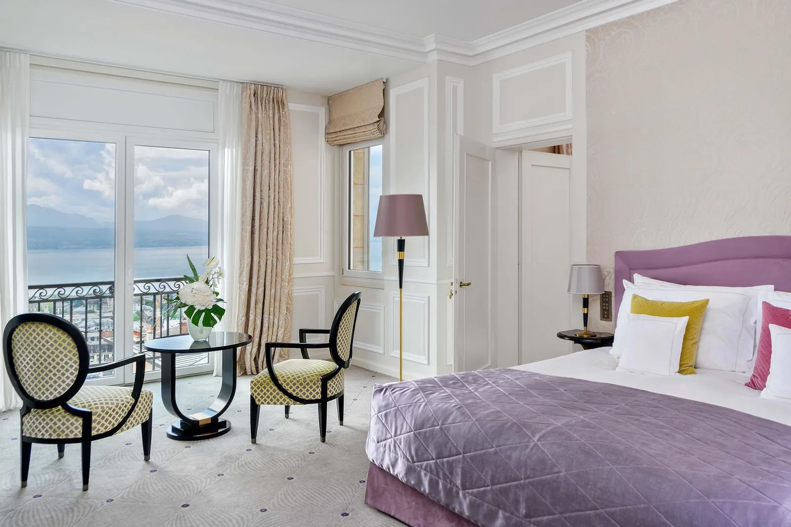 A Suite Lake View: a bed with a lilac bedspread, 2 chairs and a round table by the French window onto the balcony, with the lake and mountains