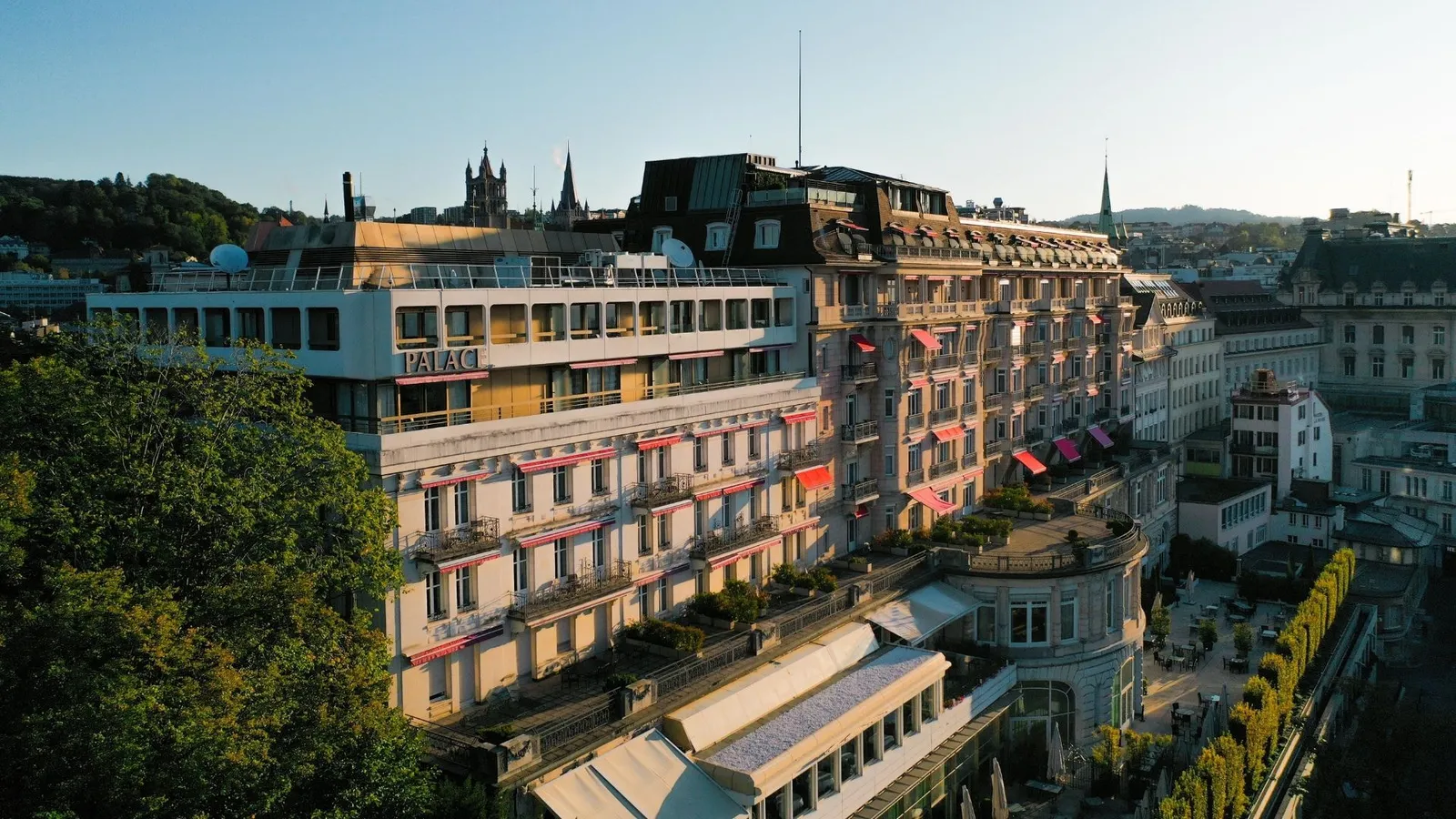 Lausanne Palace from above in late-afternoon light: the Belle Époque wings with red awnings, the modern top floor, the corner rotunda and the hedged terrace on the lake side; the cathedral towers behind