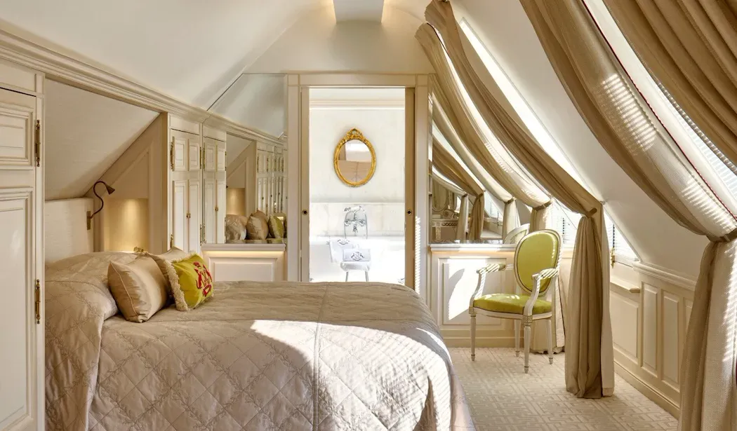 La Parisienne suite at Le Meurice, with a bed beneath a sloping ceiling, draped curtains and a green chair beside the window