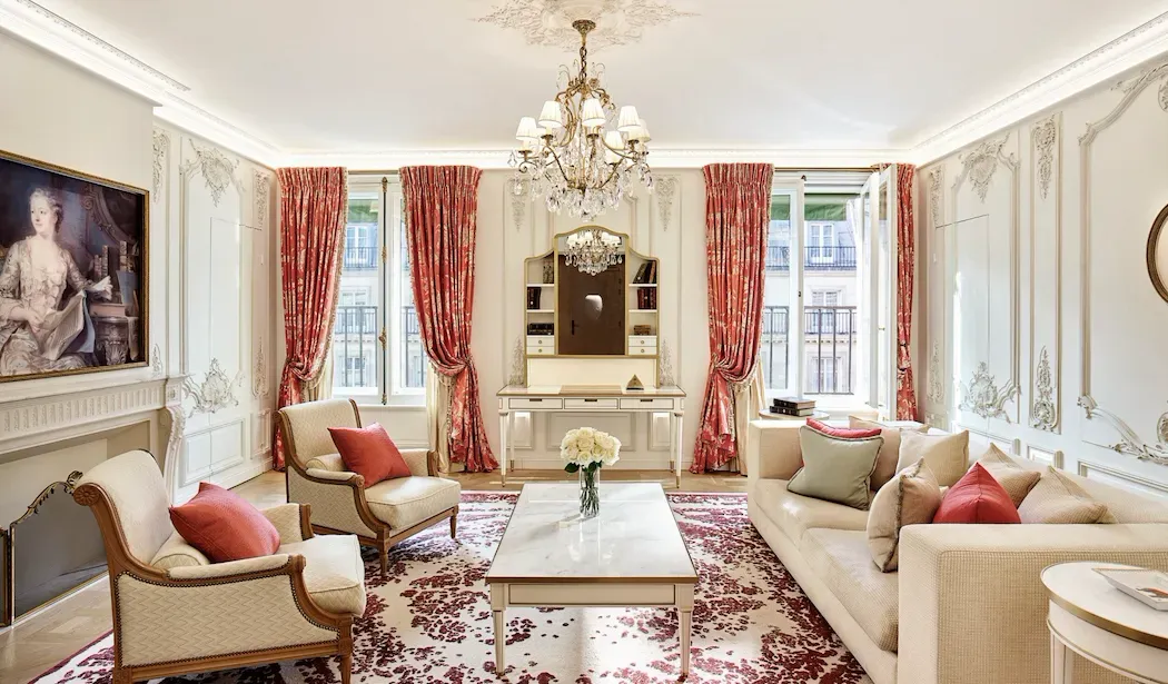 Pompadour Suite at Le Meurice, with a chandelier, cream sofas and armchairs, and red curtains at tall windows