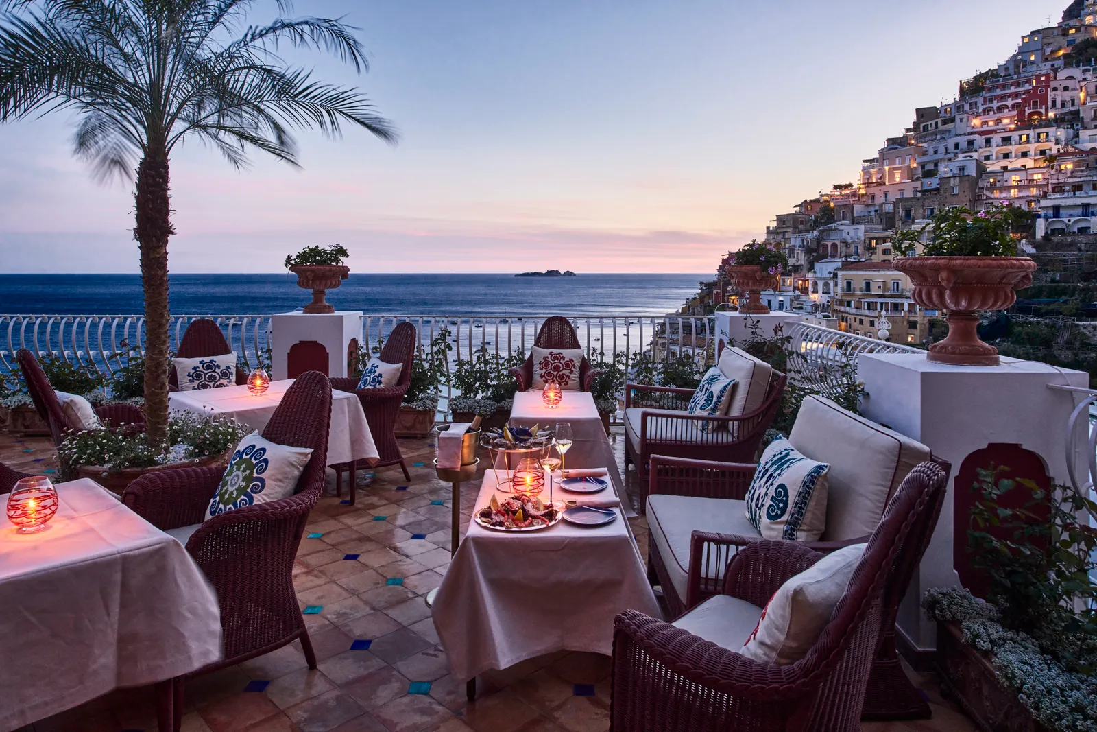 Aldo’s terrace at dusk: candlelit tables, wicker armchairs, a palm and the houses of Positano above the sea.