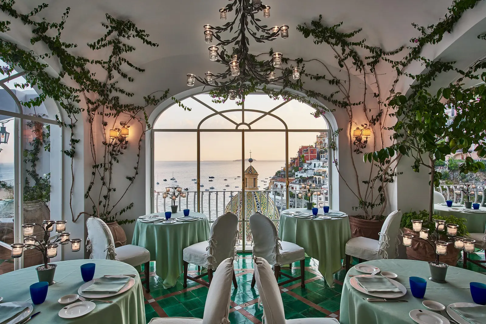 La Sponda by day: tables in green cloths, the chandelier among the climbing plants and the great arched window onto the village.