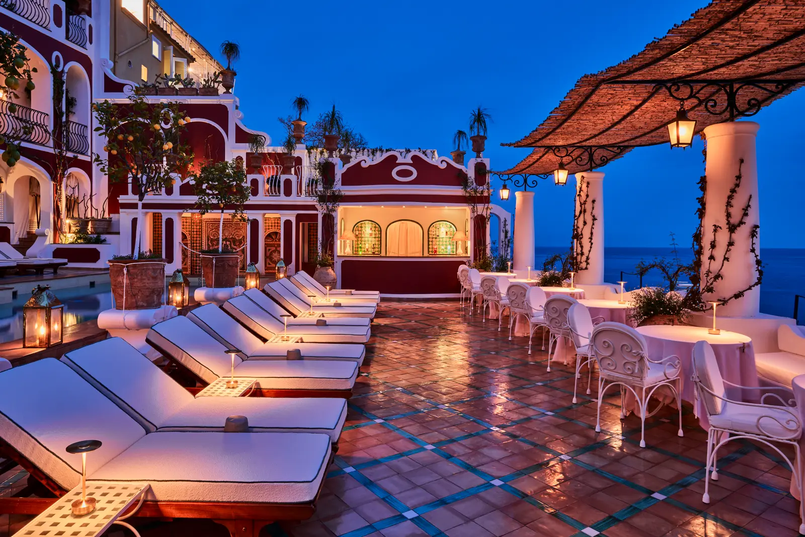 The pool terrace at dusk, the white loungers in a row, the lanterns lit and the Pool Bar counter under the pergola.