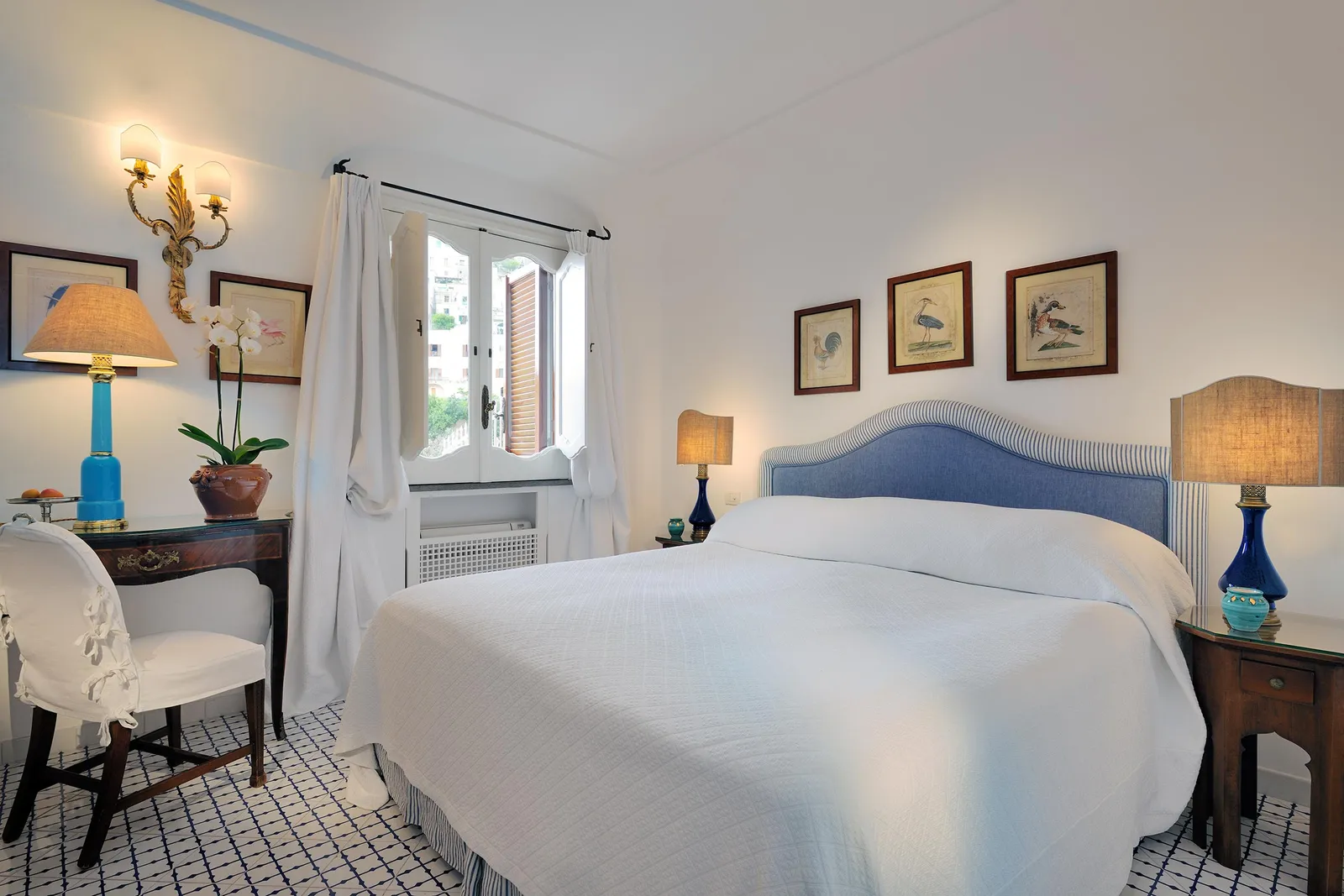 A Courtyard Room: the bed with its blue headboard, blue glass lamps, bird prints and the window onto the courtyard.