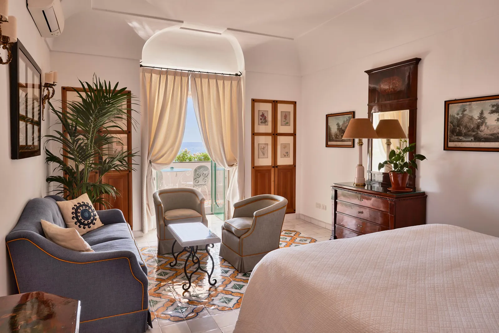 A Deluxe Sea View room: the blue sofa, the armchairs, a potted palm and the French window open to the sea.