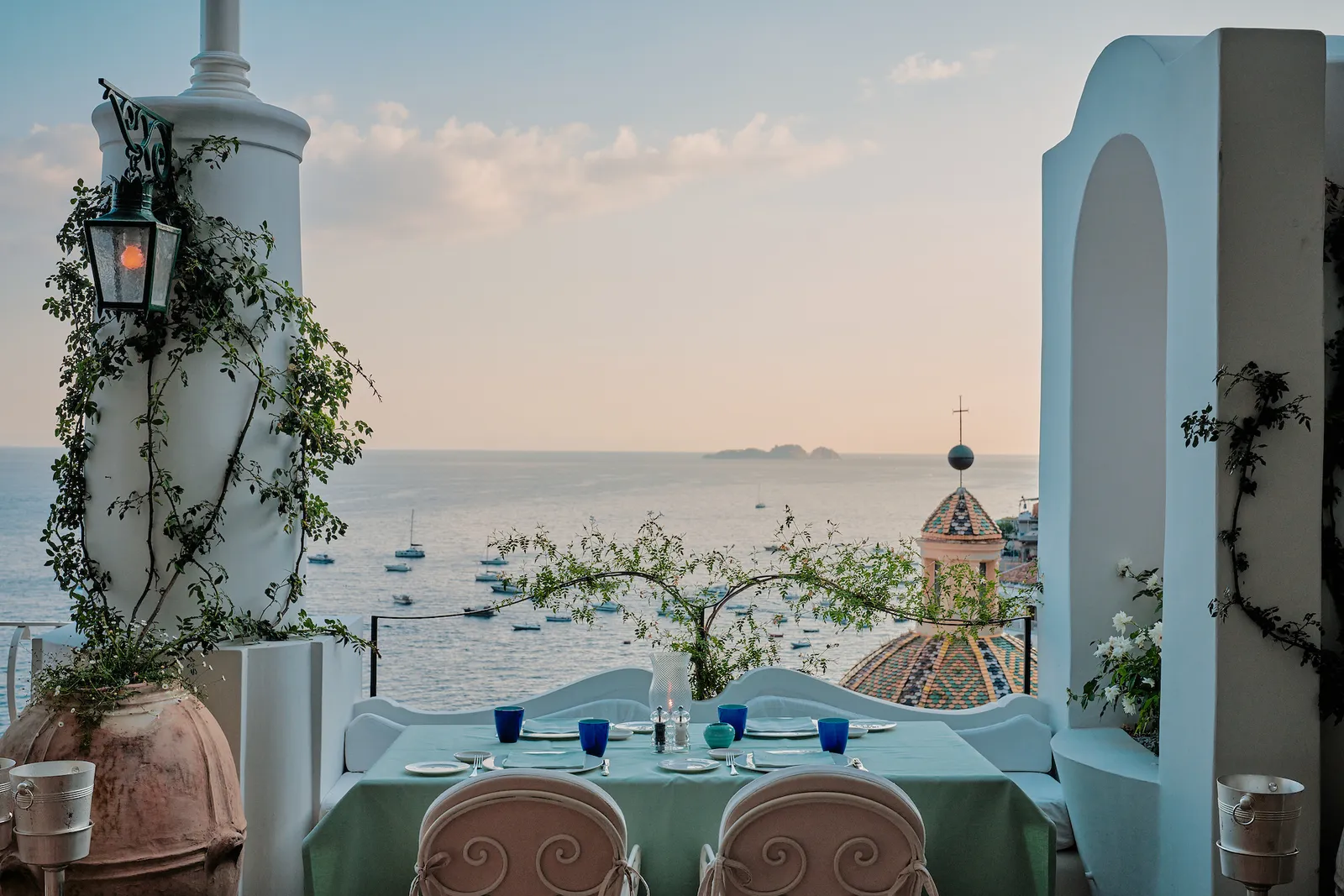 A table laid on the terrace between 2 white columns, with the majolica dome of Santa Maria Assunta and the sea.