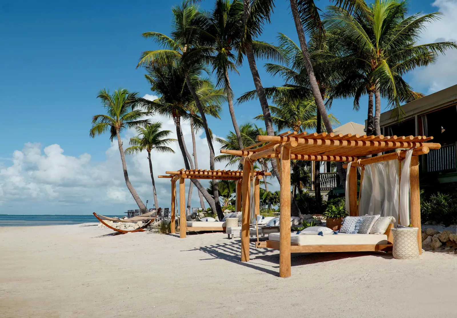 2 timber pergolas with beach beds on white sand, a hammock and coconut palms.