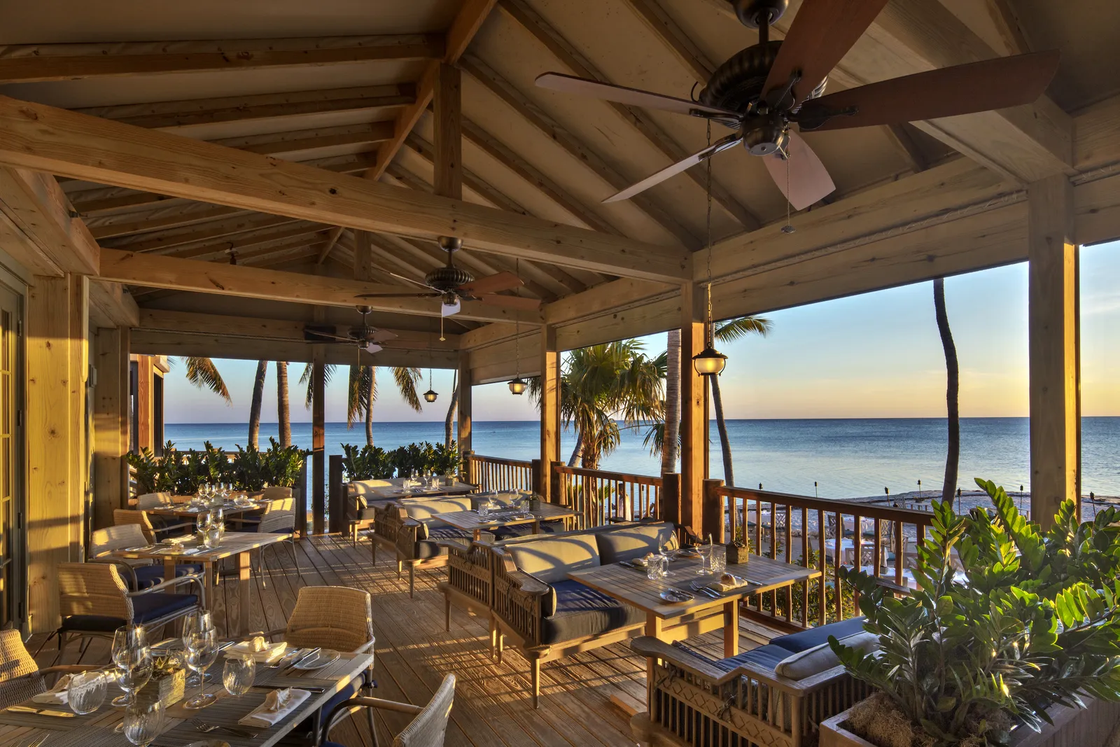 The Dining Room’s verandah under its timber roof and ceiling fans: tables laid, and the ocean at sunset beyond the rail.