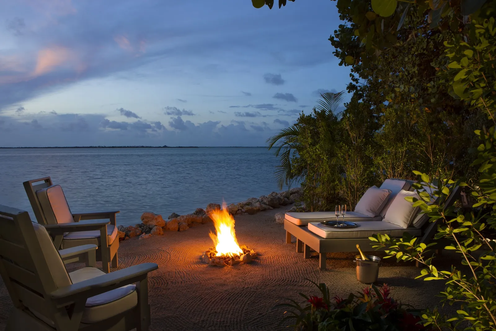 A firepit alight on the sand at dusk, between 2 armchairs and a lounger, the calm sea in front.