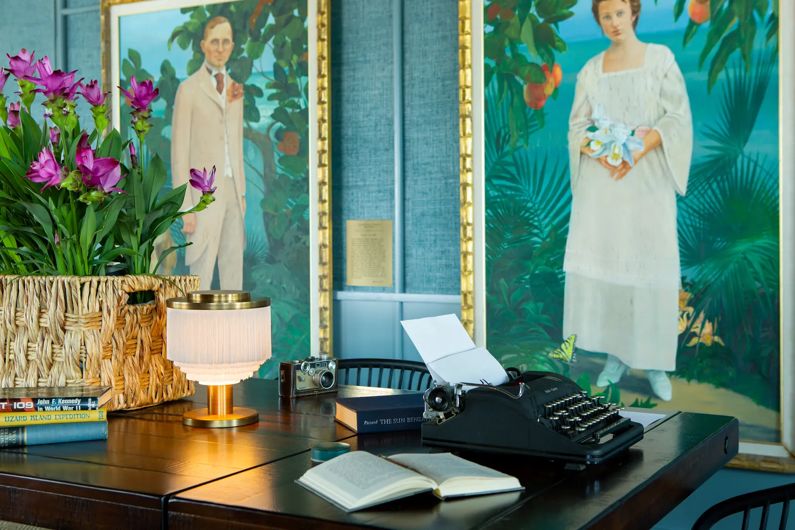 A corner of the Great Room: a typewriter on a desk, orchids and 2 painted full-length portraits.