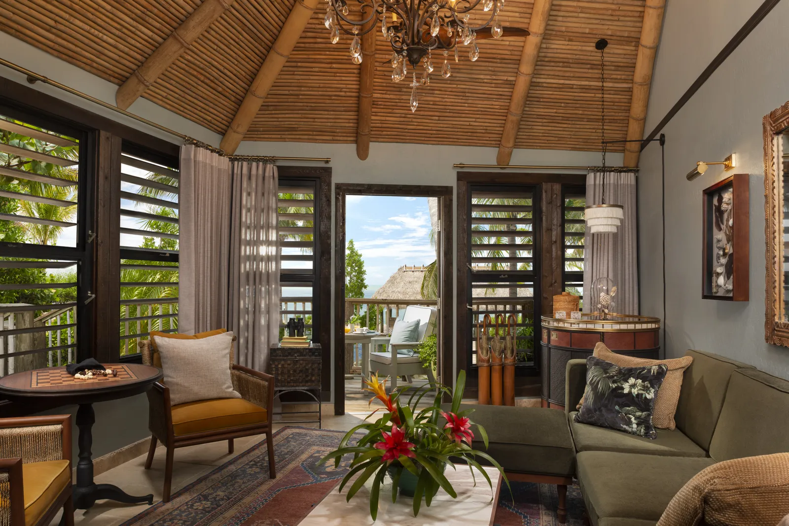 An Island Escape Suite’s living room: the bamboo ceiling and chandelier, a green sofa, louvred shutters and the door open to the deck.
