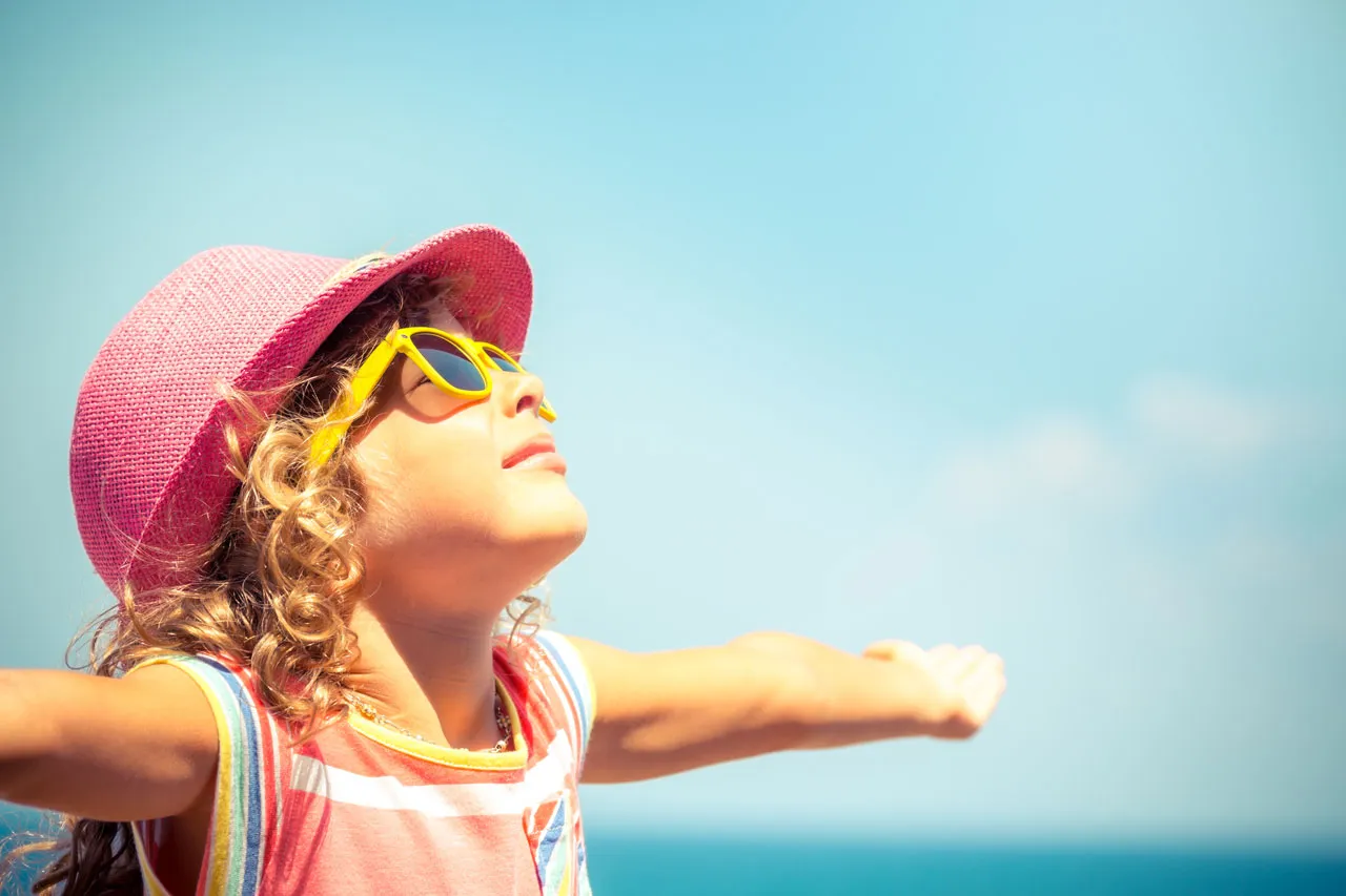 A little girl in a sunhat and sunglasses, arms open to the sky