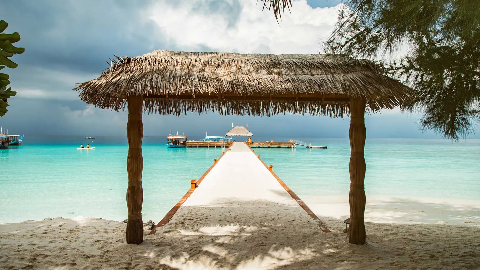 A thatched shelter on the beach, with the jetty and the arrivals platform on the lagoon behind it.