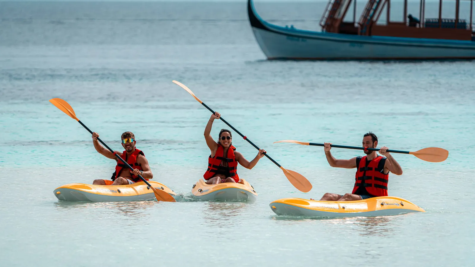 3 guests paddling canoes in the shallow lagoon, with a Maldivian boat behind.