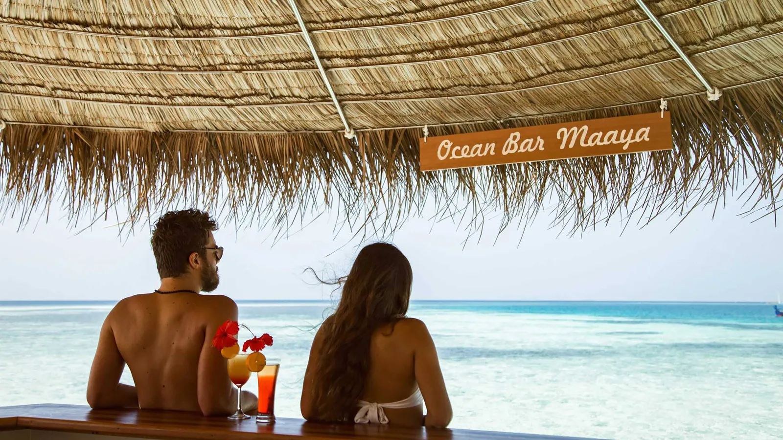 A couple seen from behind at the Ocean Bar Maaya counter, 2 cocktails and the lagoon before them.