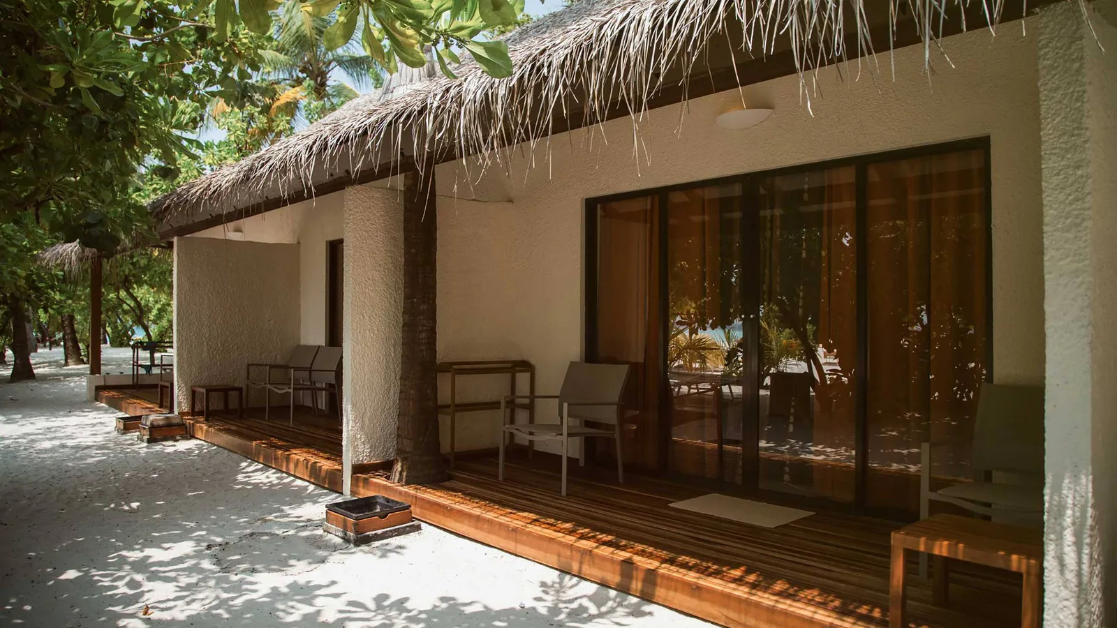 A Beach Bungalow with its thatched roof and timber veranda on the sand, among the trees.