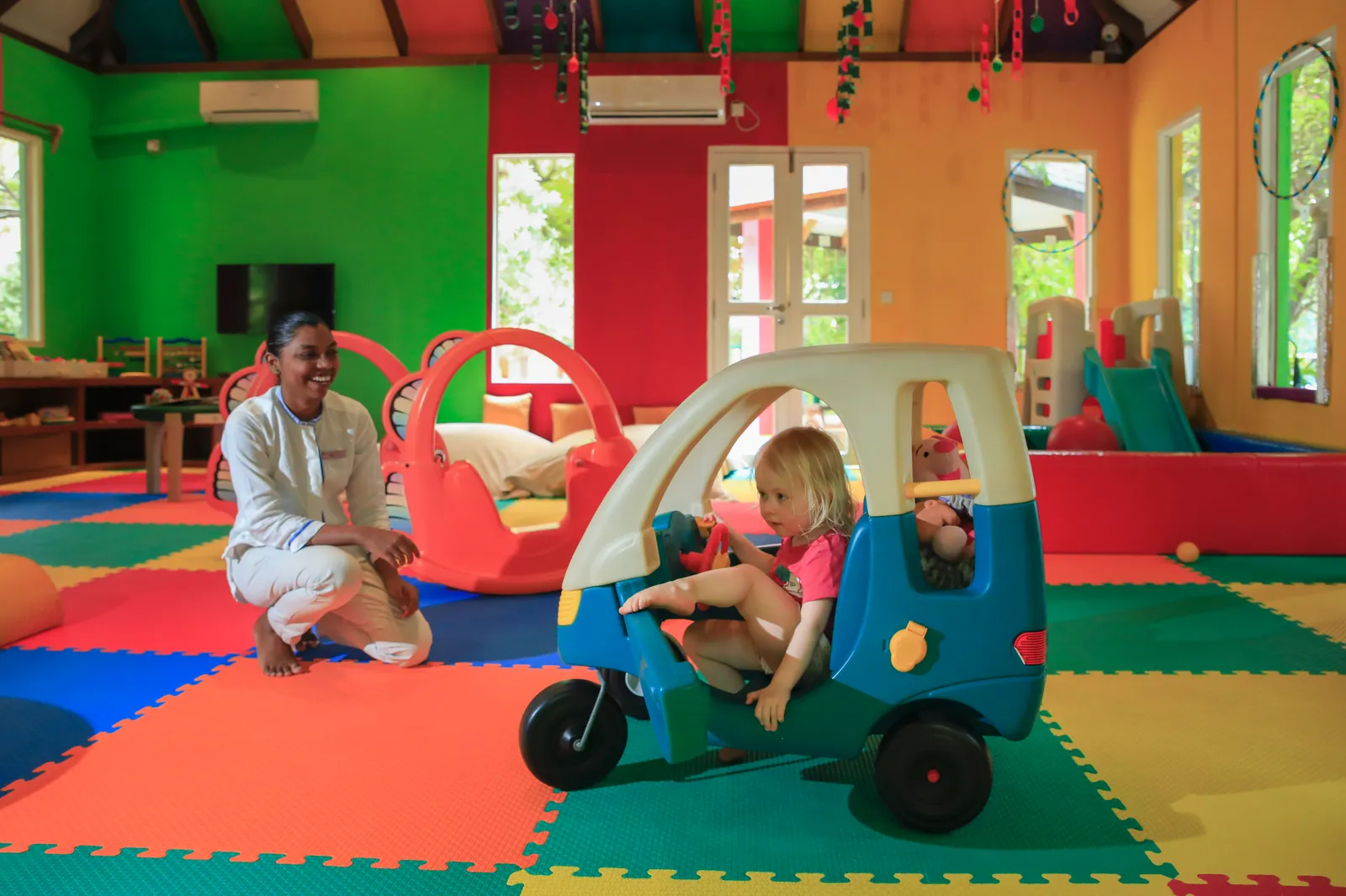 A little girl in a pedal car in Camp Safari’s brightly painted room, beside a kneeling member of staff.