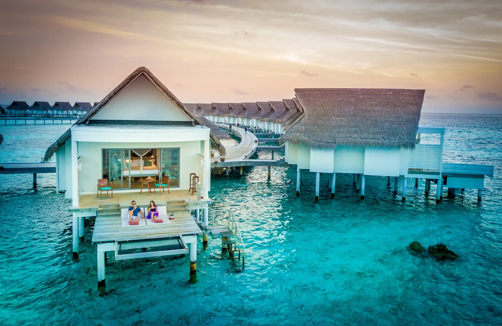 A water villa at sunset, 2 guests on its deck, the steps down to the lagoon and the row of villas behind.