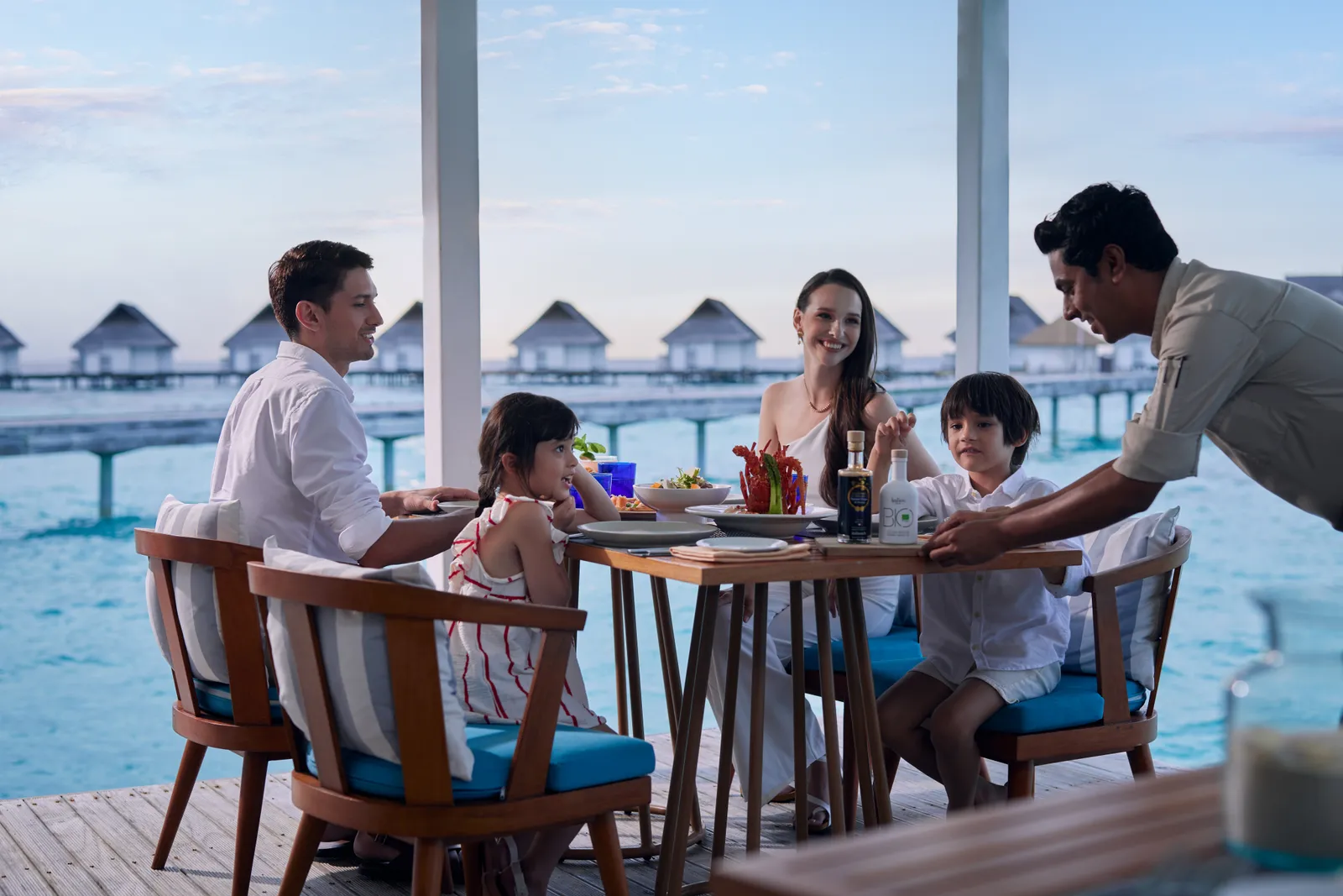 A family at table on Mare Azzurro’s terrace, served by a waiter, with the water villas behind.
