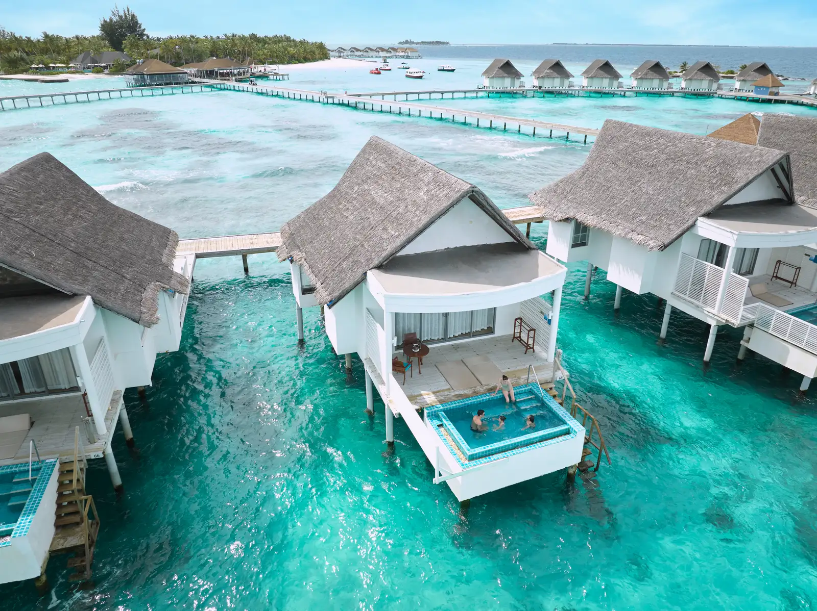 The Club Sunset Overwater Pool Villas from above: a deck with its small infinity pool over the lagoon and 3 guests in the water.