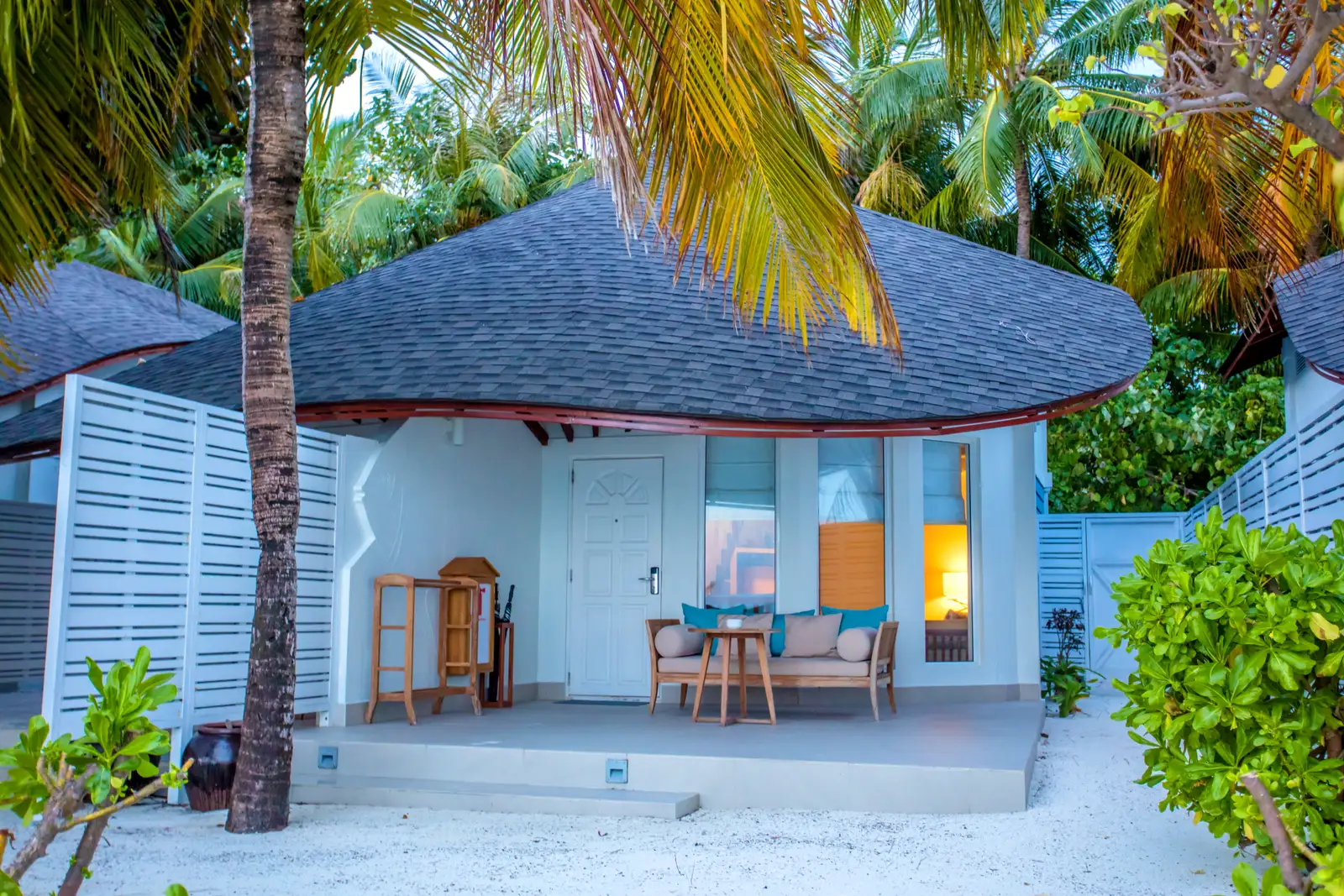 The Duplex Beach Villa from outside: the white cottage with its conical roof, the veranda and the palms on the sand.