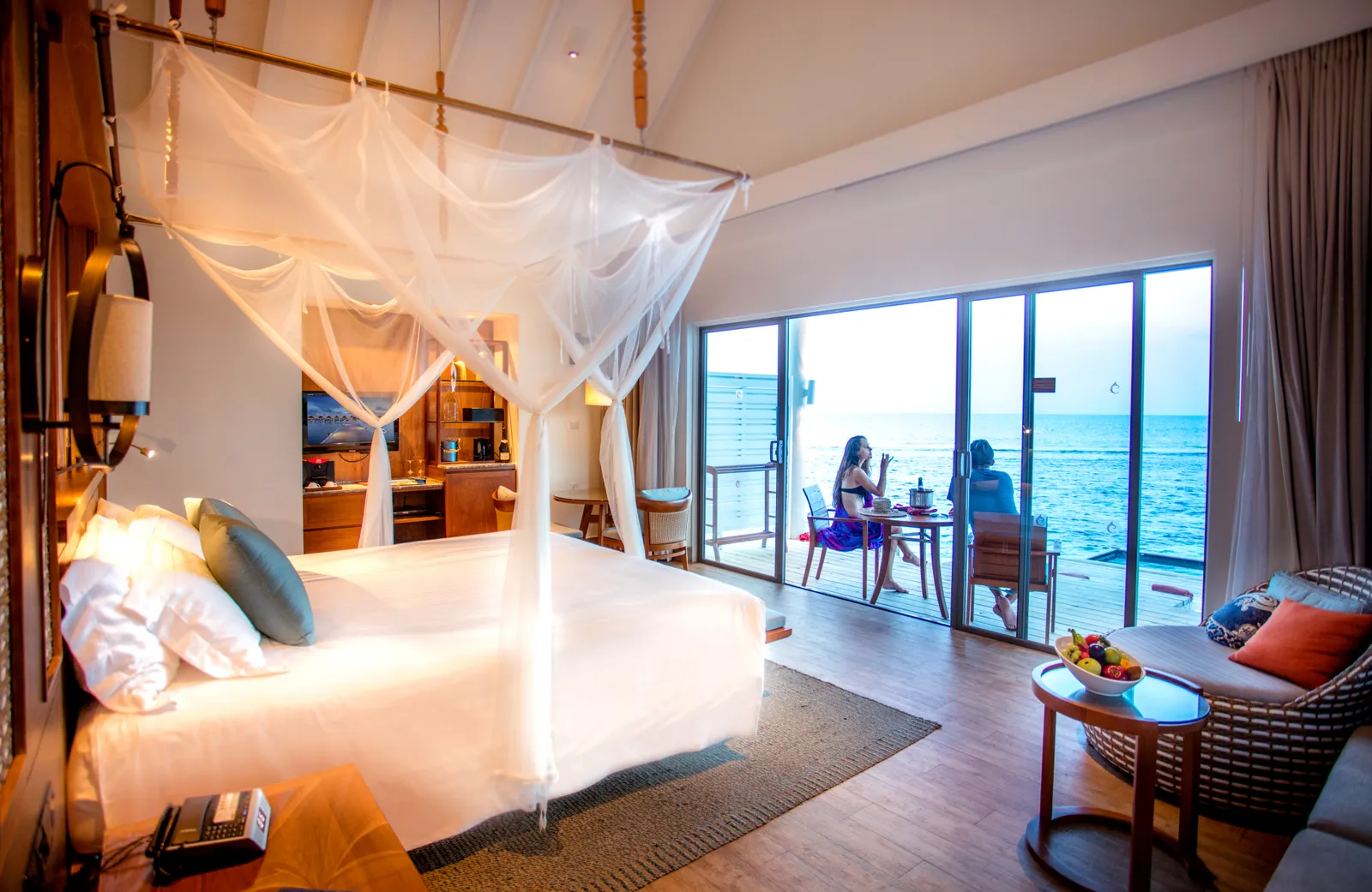 A Reethi Muraka Overwater Villa bedroom: the bed under its mosquito net and, beyond the glass, 2 guests at table on the deck.