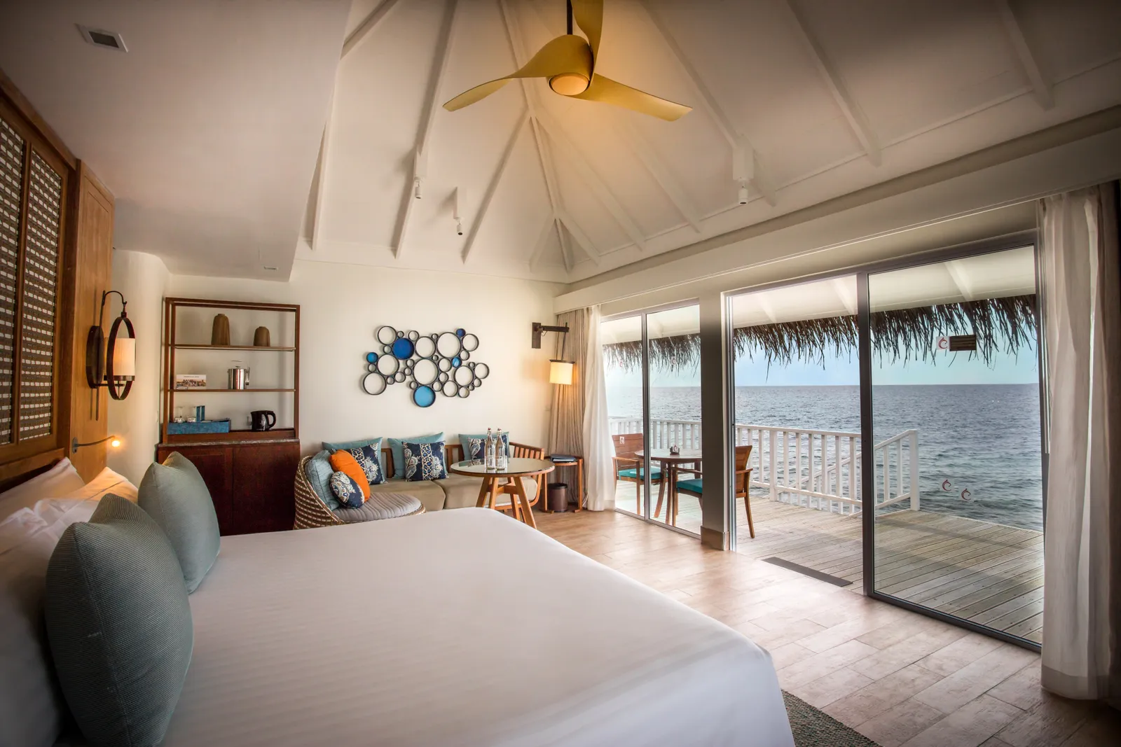 A Sunrise Overwater Villa bedroom: the bed, the sitting corner and the glass doors open to the deck and the sea.
