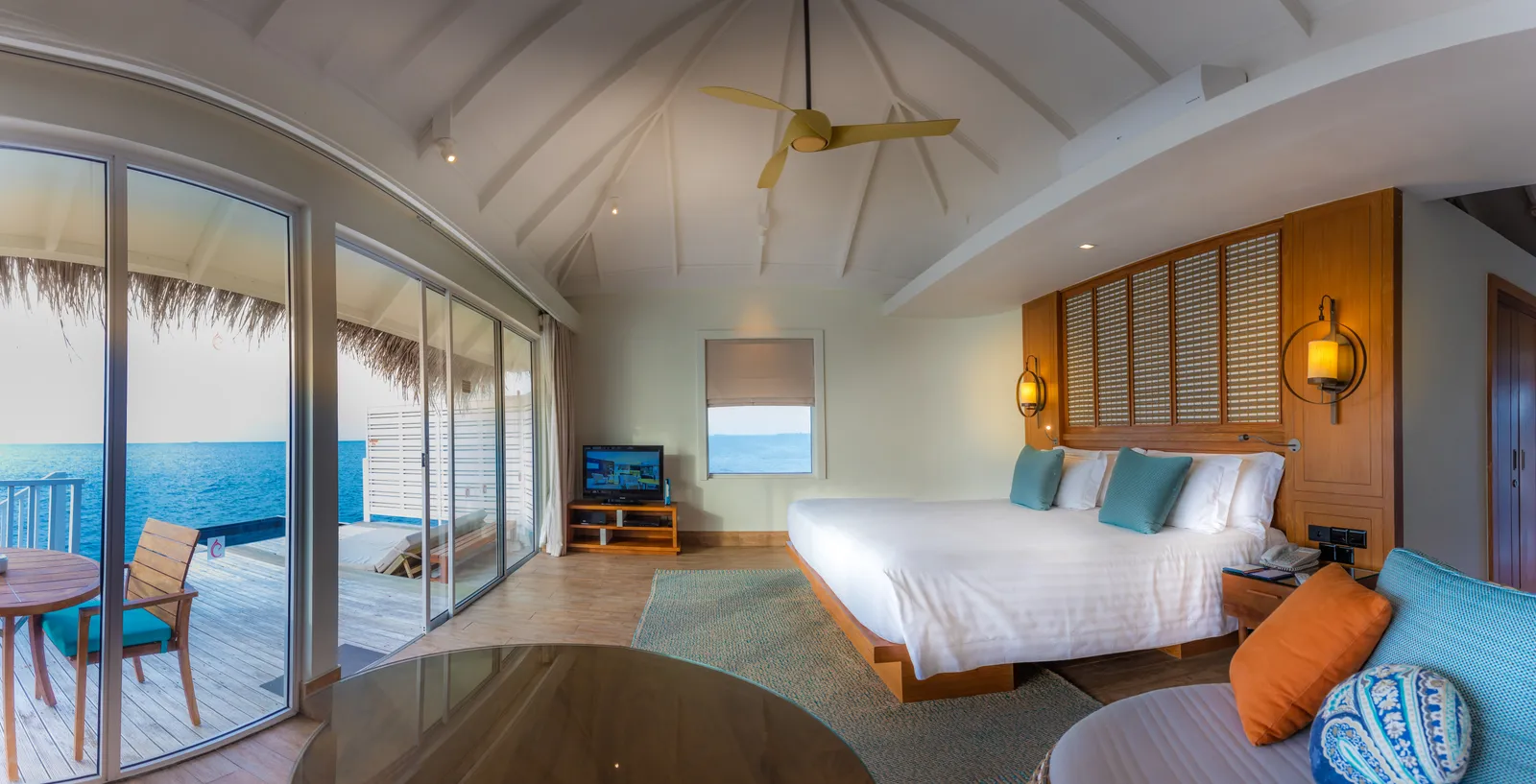 A Sunset Overwater Villa bedroom, with the king bed, the television and the deck and its lounger beyond the glass.
