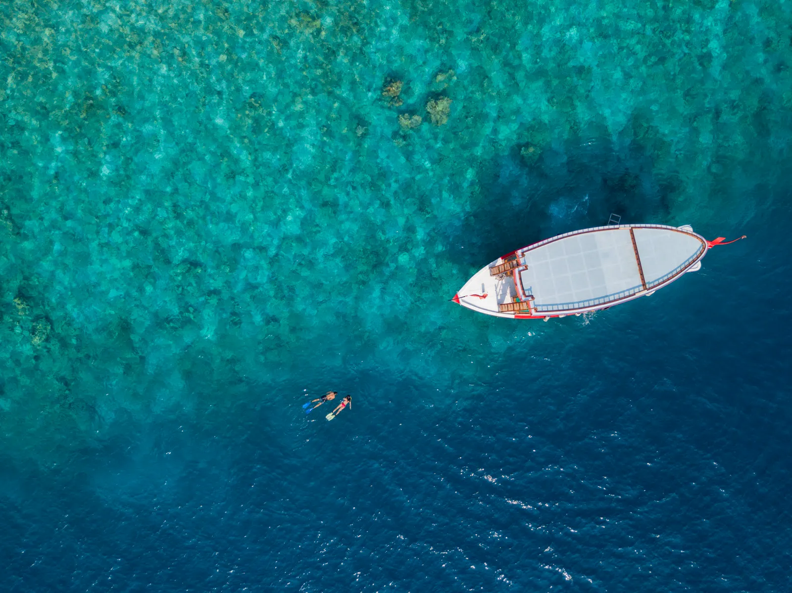 A white dhoni seen from above over the reef, with 2 people snorkelling beside it.