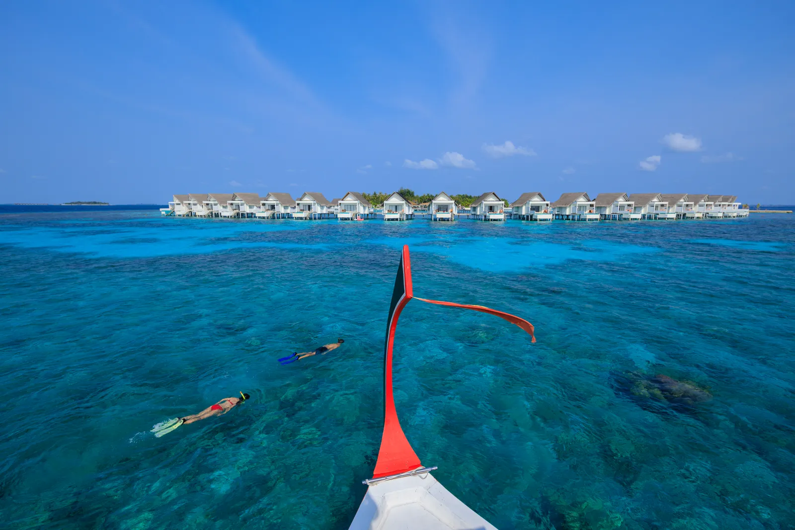 The prow of a dhoni over the reef, 2 people snorkelling and the row of water villas on the horizon.