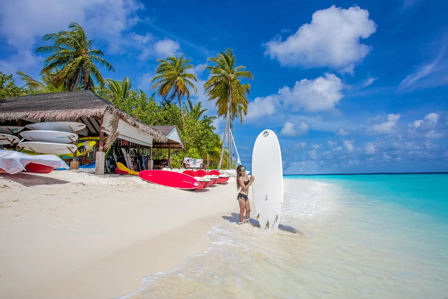 The water sports beach, kayaks and boards on the sand and a young woman holding a surfboard at the water’s edge.