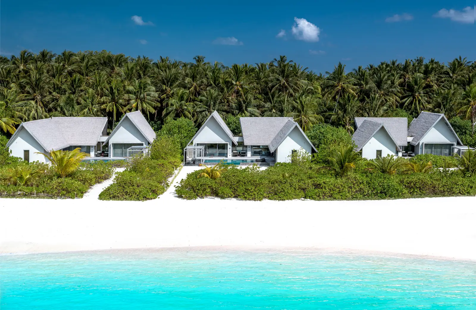 The white-roofed beach villas in a row among the palms, with the sand and the lagoon in front.